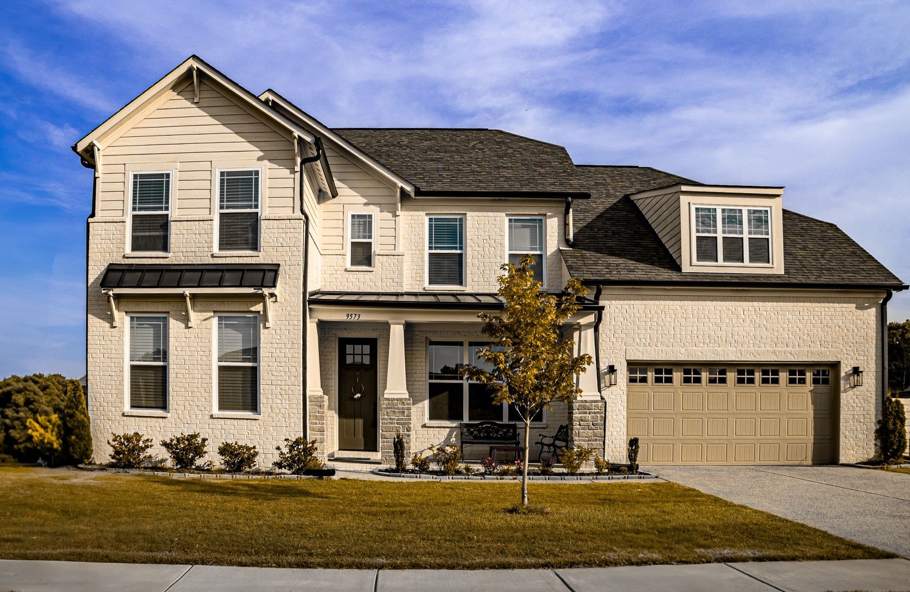 Two-story beige brick suburban house with a two-car garage and small front yard.