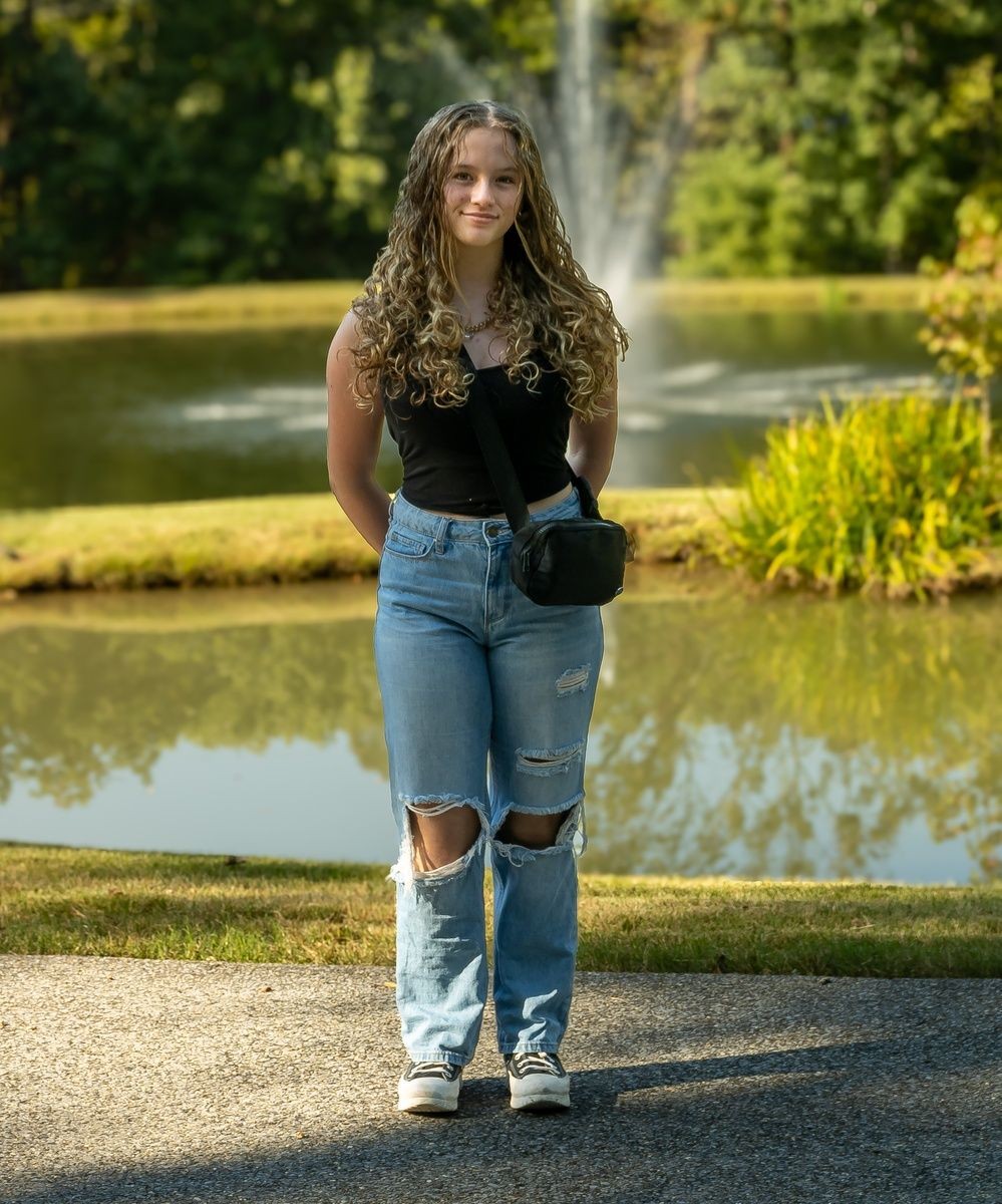 Young woman with curly hair in casual attire standing by a pond with a fountain and greenery.